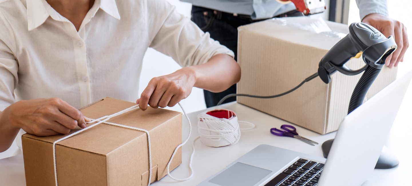Duas pessoas embalam caixas com laptop, scanner, tesoura e outros itens sobre a mesa.