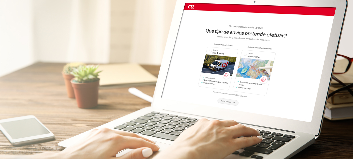 Pessoa a usar um portátil com uma página sobre tipos de envios, ao lado de um telemóvel e uma planta.