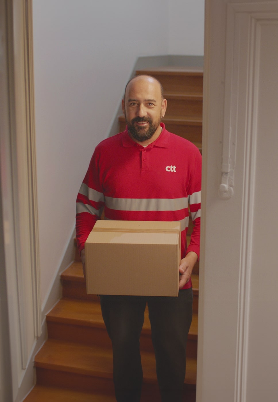 Duas pessoas numa sala, uma está sentada ao computador com livros e papéis, outra está com a farda dos CTT e com uma encomenda na mão.